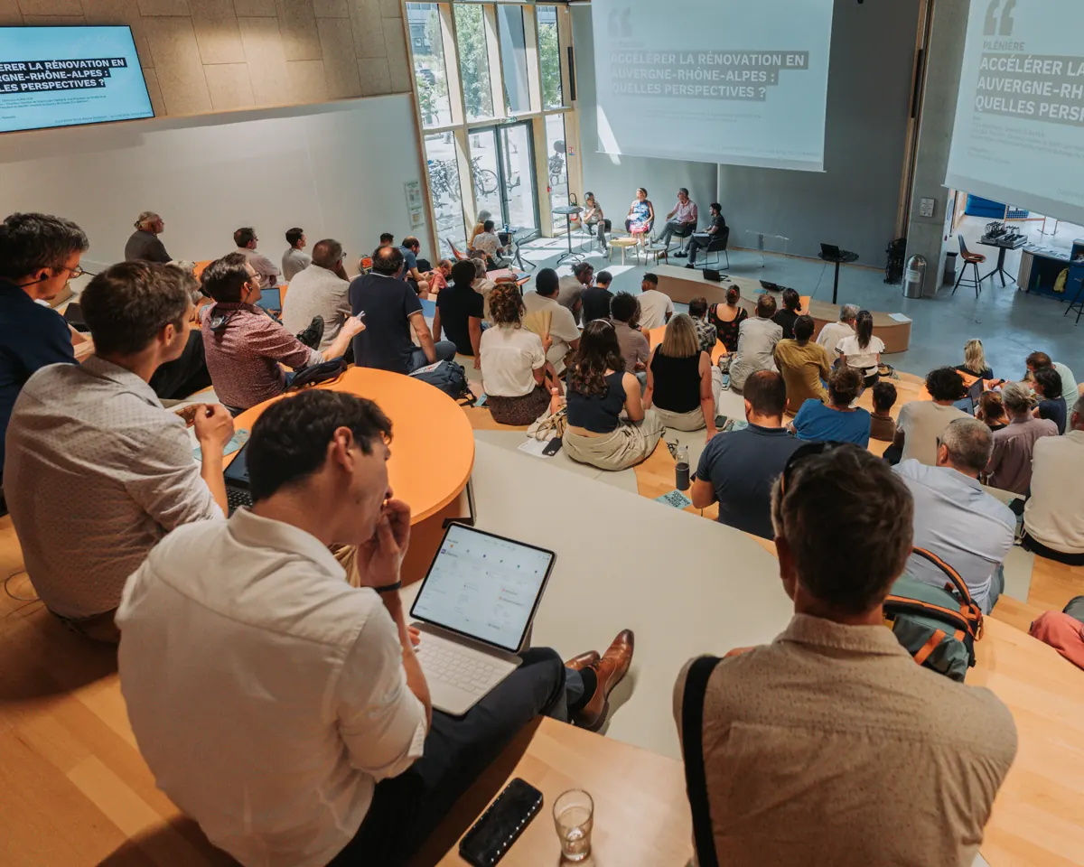 Salle de conférence avec écrans de projection allumés et public assis, vue depuis le fond