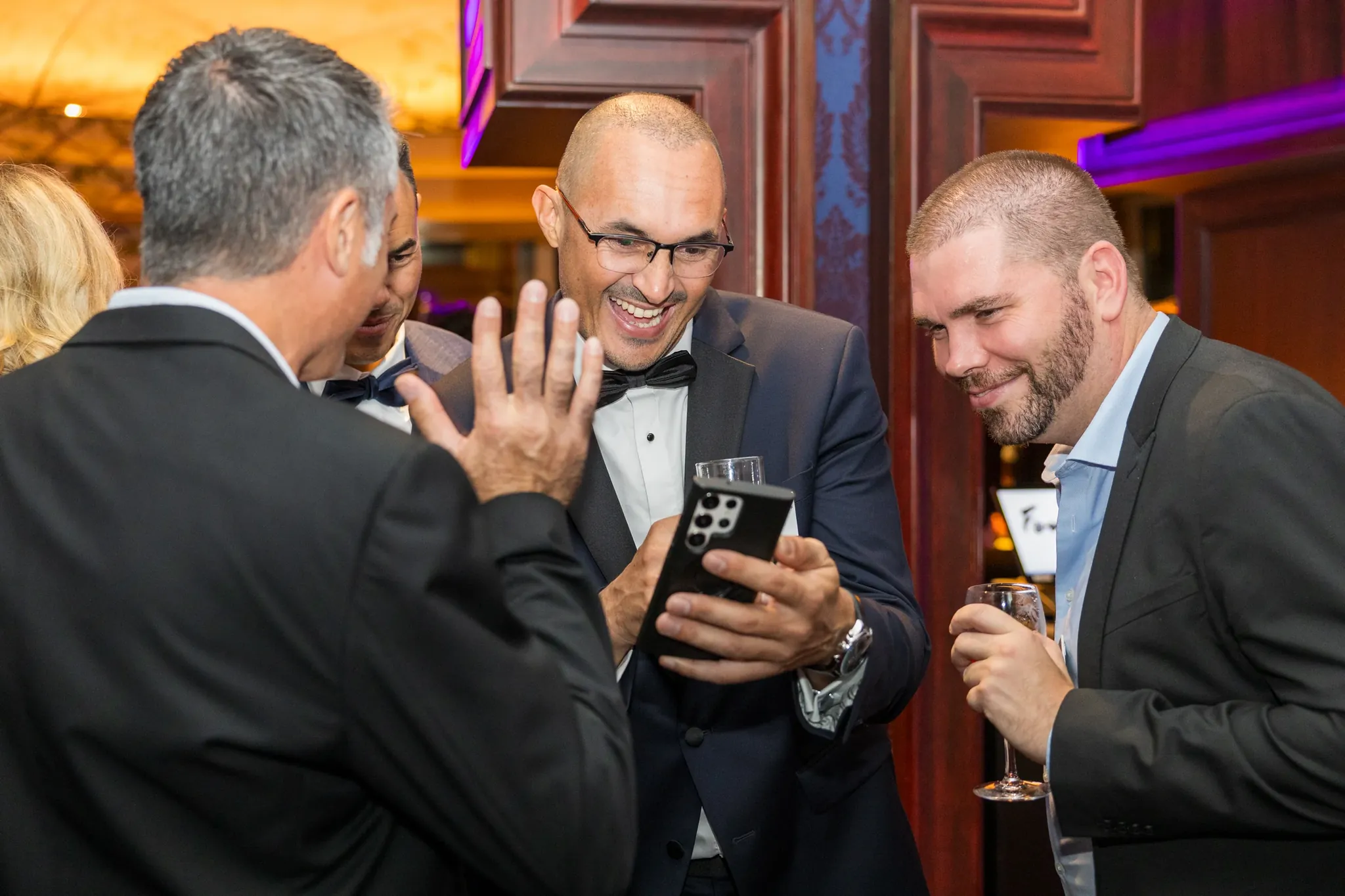 Invités en tenue de soirée qui rient autour d'un téléphone lors d'un dîner de gala, ambiance chaude et colorée préservée