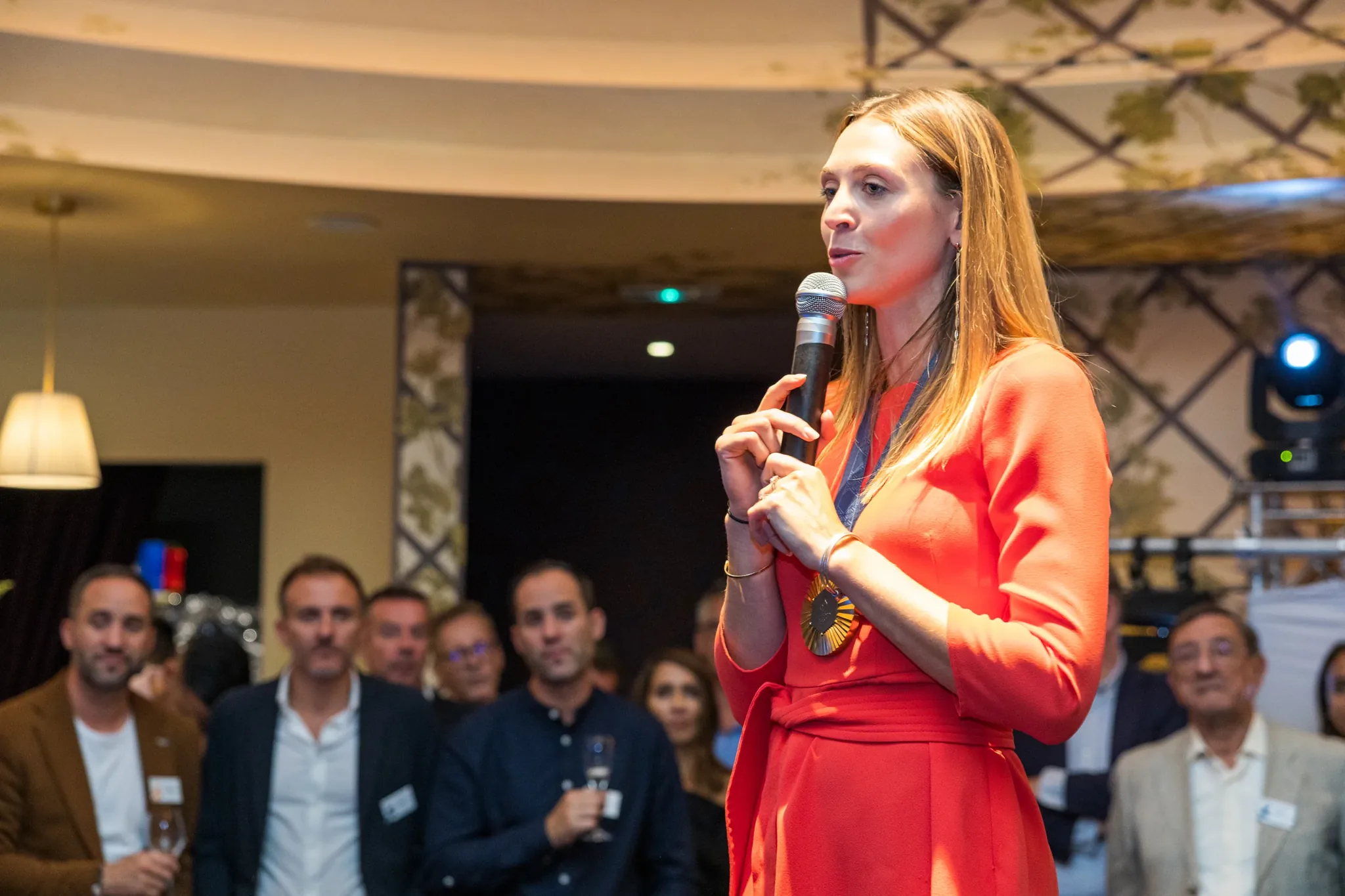 Intervenante en robe au micro lors d'une soirée d'entreprise, menton légèrement baissé, mains occupées, pose naturelle en prise de parole