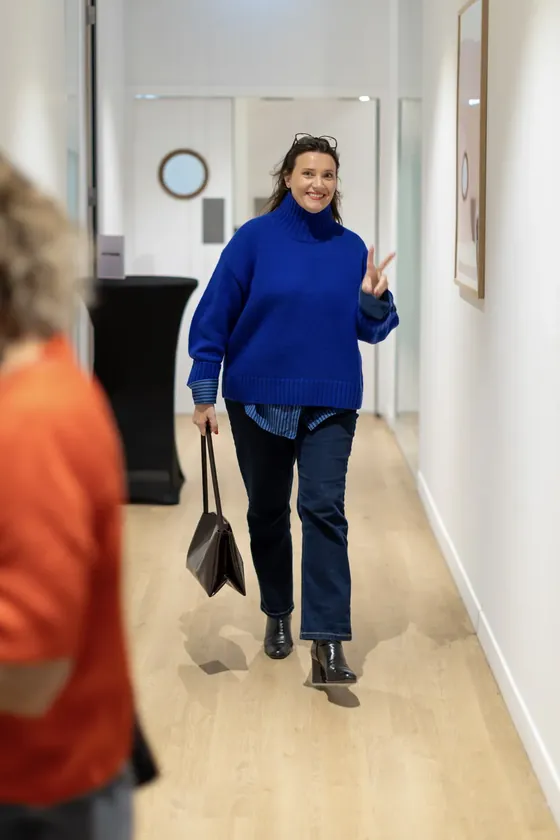 Une femme en pull bleu vif marche dans un couloir clair en parquet, sac à la main, et lève la main en souriant