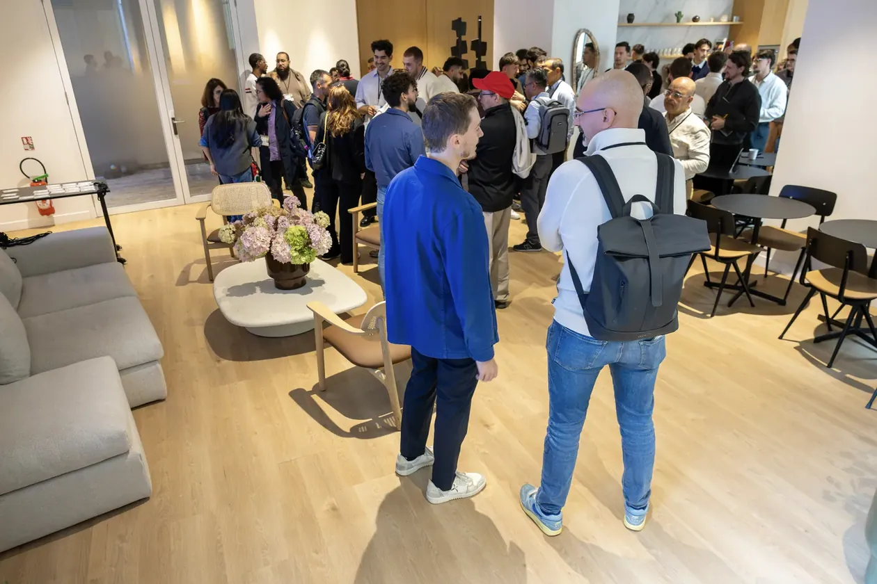 Vue large du salon avec une foule dense au fond et deux hommes de dos au premier plan, dont un sac à dos, parquet clair