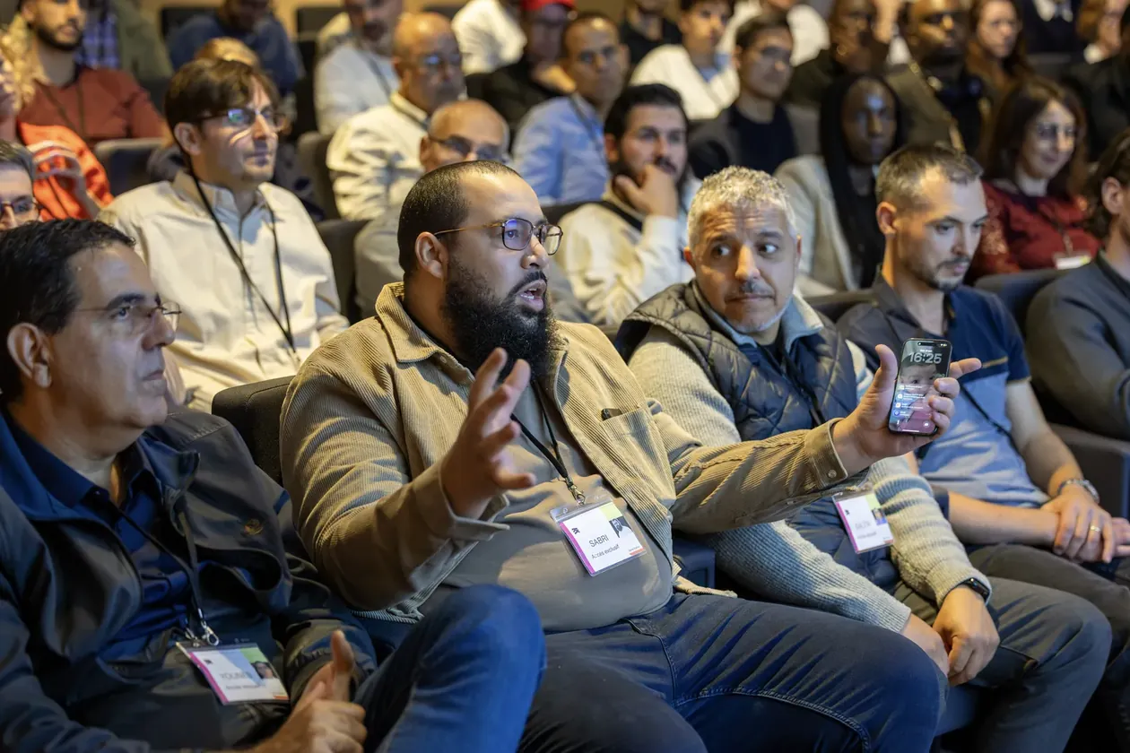 Un homme barbu en veste beige parle en gesticulant dans le public, smartphone en main, voisins attentifs, badges visibles
