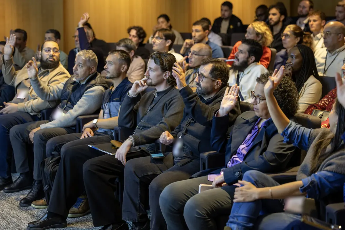 Salle de conférence aux murs de bois : des dizaines de participants assis en rangs lèvent la main, certains très haut ; d'autres écoutent, la main près du visage