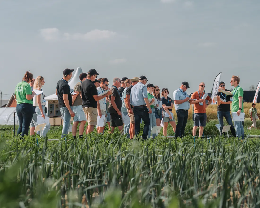 Groupe de participants debout dans un champ cultivé pendant une visite en plein jour, badge au cou et documents en main, tente blanche et oriflammes au fond