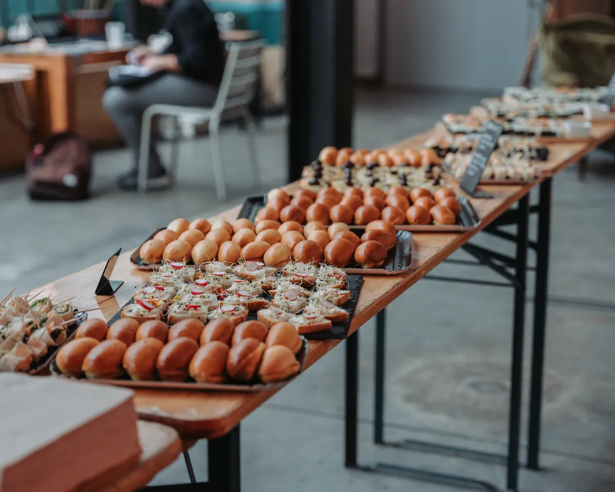 Long buffet dressé sur des tables en bois à pieds noirs : plateaux de petits pains briochés dorés, bouchées garnies de radis, petites ardoises ; au fond, dans le flou, une personne assise consulte des papiers